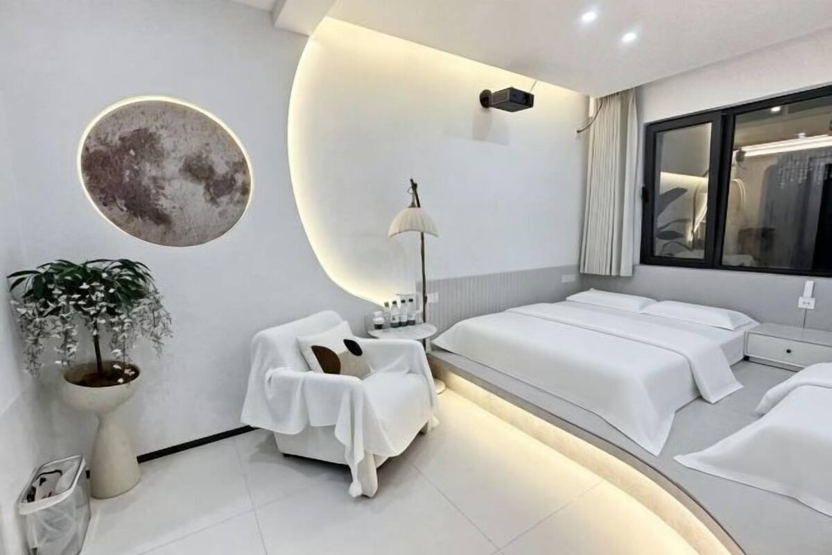 A modern hotel room with a large bed, a seating area, a bedside table, a lamp, a potted plant, and a large window.