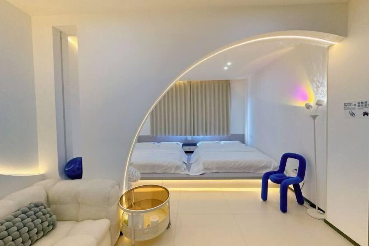 A modern hotel room with a curved ceiling, a bed with white linens, a small seating area with a white sofa, a blue chair, and a floor lamp.