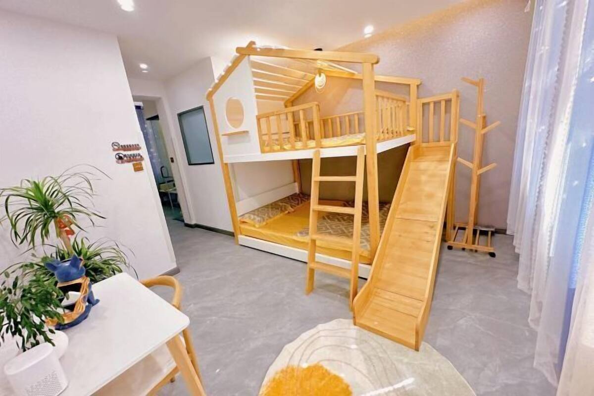 A bunk bed with a slide, a small table with a plant, and a window with curtains.