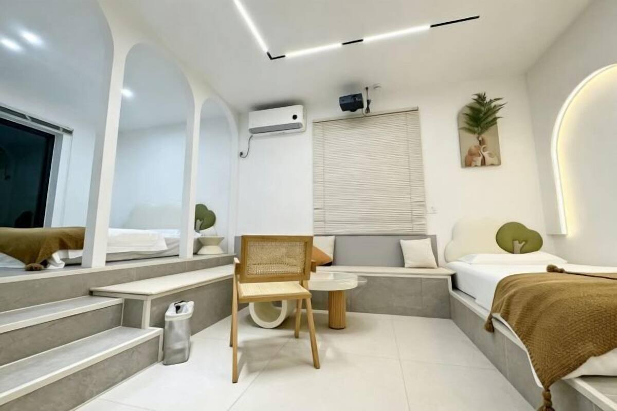 A modern hotel room with a sofa, a chair, a small table, a bed, a window with blinds, a wall-mounted air conditioner, and a ceiling light fixture.