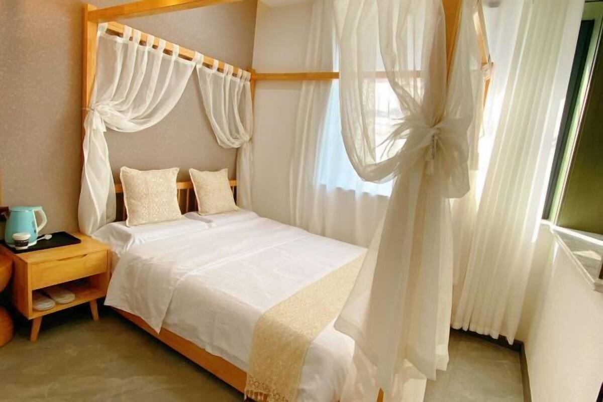 A bed with a canopy, a nightstand with a kettle, and a window with curtains.