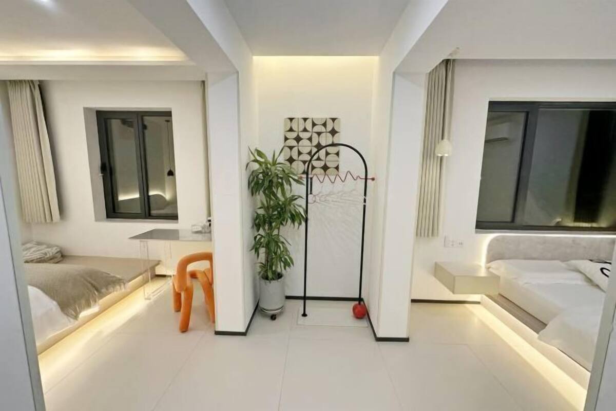 A modern hotel room with a bed, bedside table, chair, potted plant, and a decorative wall piece.