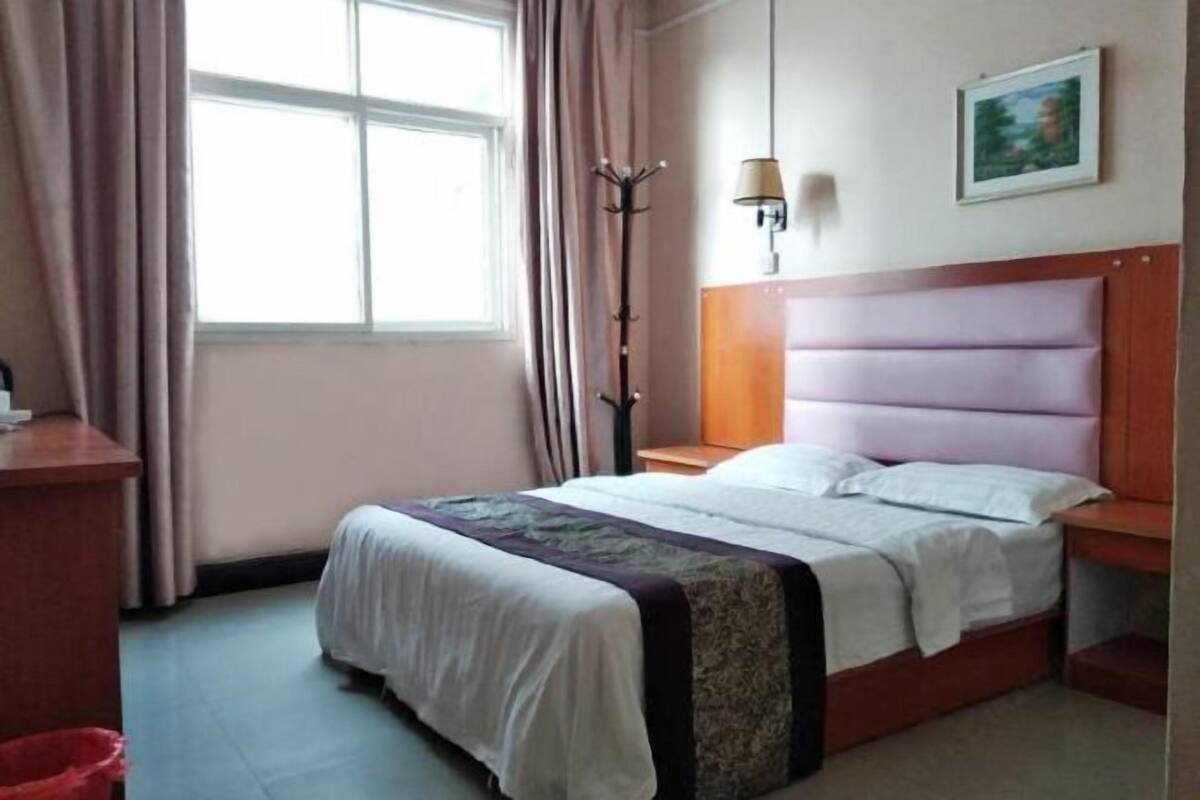 Ein Schlafzimmer mit einem Bett, einem Nachttisch, einer Lampe, einem Kleiderhaken und einem Fenster mit Vorhängen.