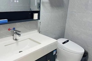 A bathroom with a white sink, a mirror, a toilet, and a shelf with a roll of toilet paper. - Minnan Lingang Mingdu Hotel (Junan County)