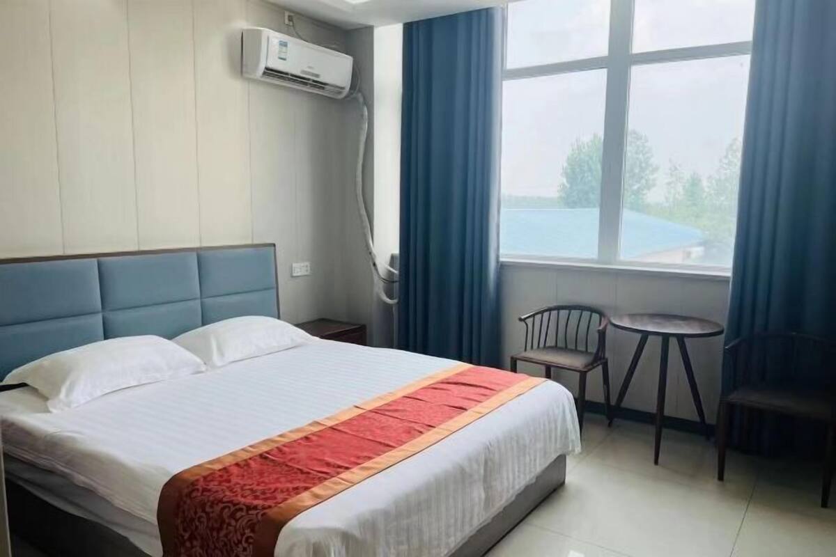 Une chambre d’hôtel comprenant un lit, une petite table, une chaise, un appareil de climatisation et une fenêtre avec des rideaux.