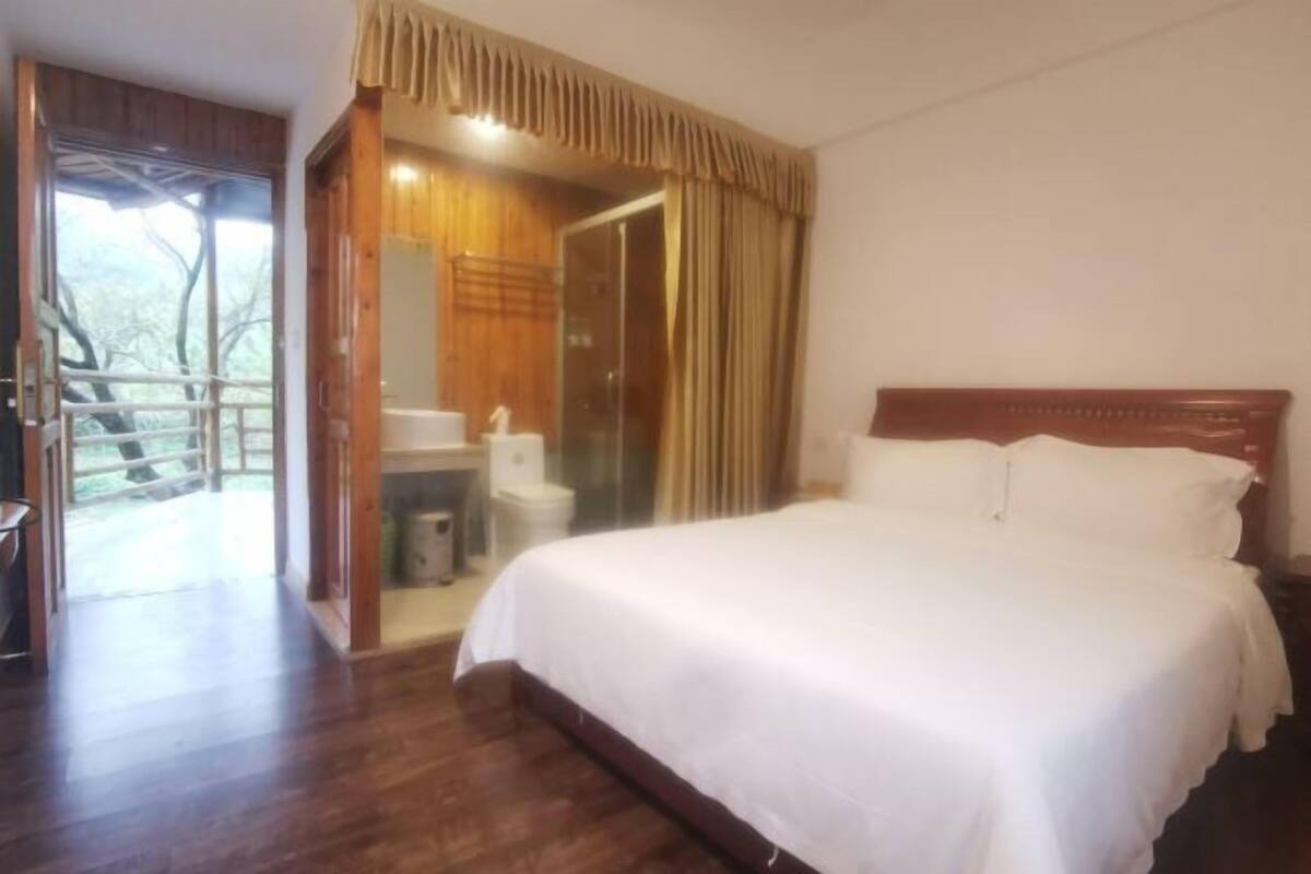 A bedroom with a large bed, wooden flooring, a shower area, and a view of trees through an open door.