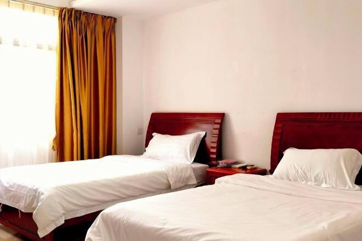 A hotel room with two single beds, wooden headboards, white bedding, a red nightstand, and a window with yellow curtains.