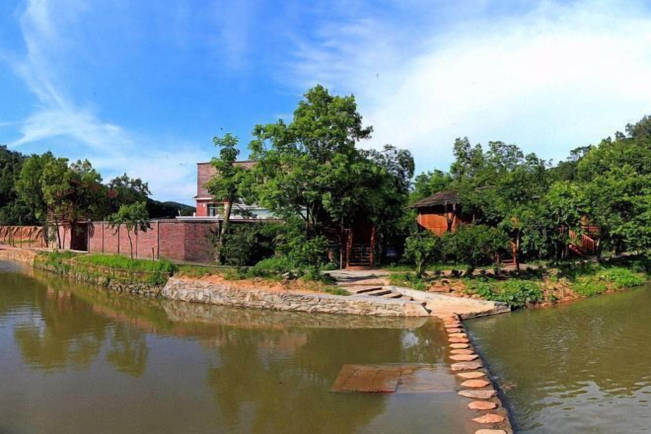 A serene riverside setting with a stone pathway leading to a bridge, surrounded by lush greenery and traditional architecture.