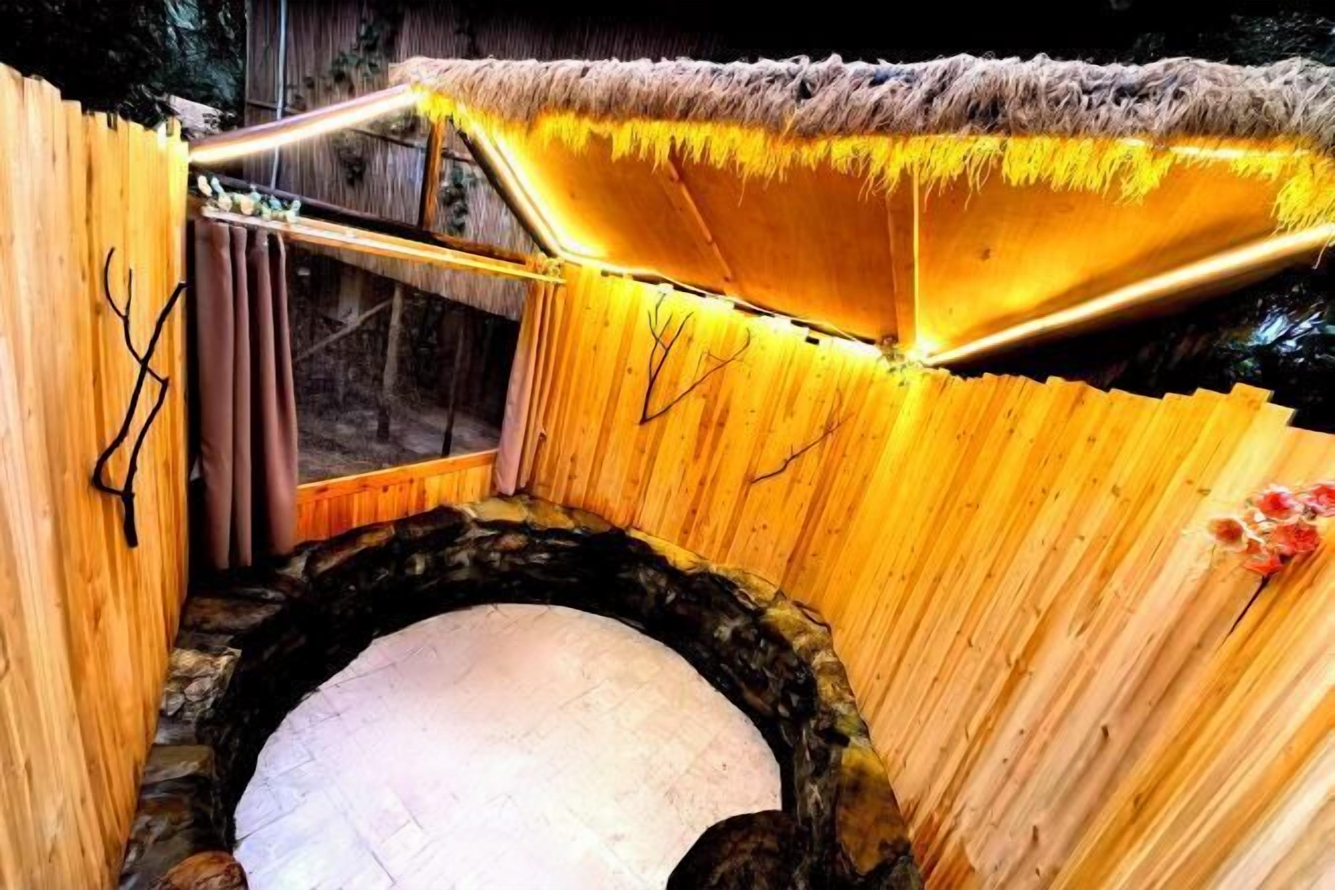 Una estructura rústica de madera con techo de paja, piso de piedra circular y banco de madera.