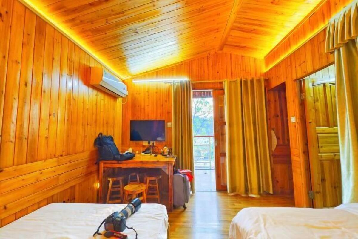 A wooden cabin room with two beds, a desk with a chair, a television, and a window with curtains.