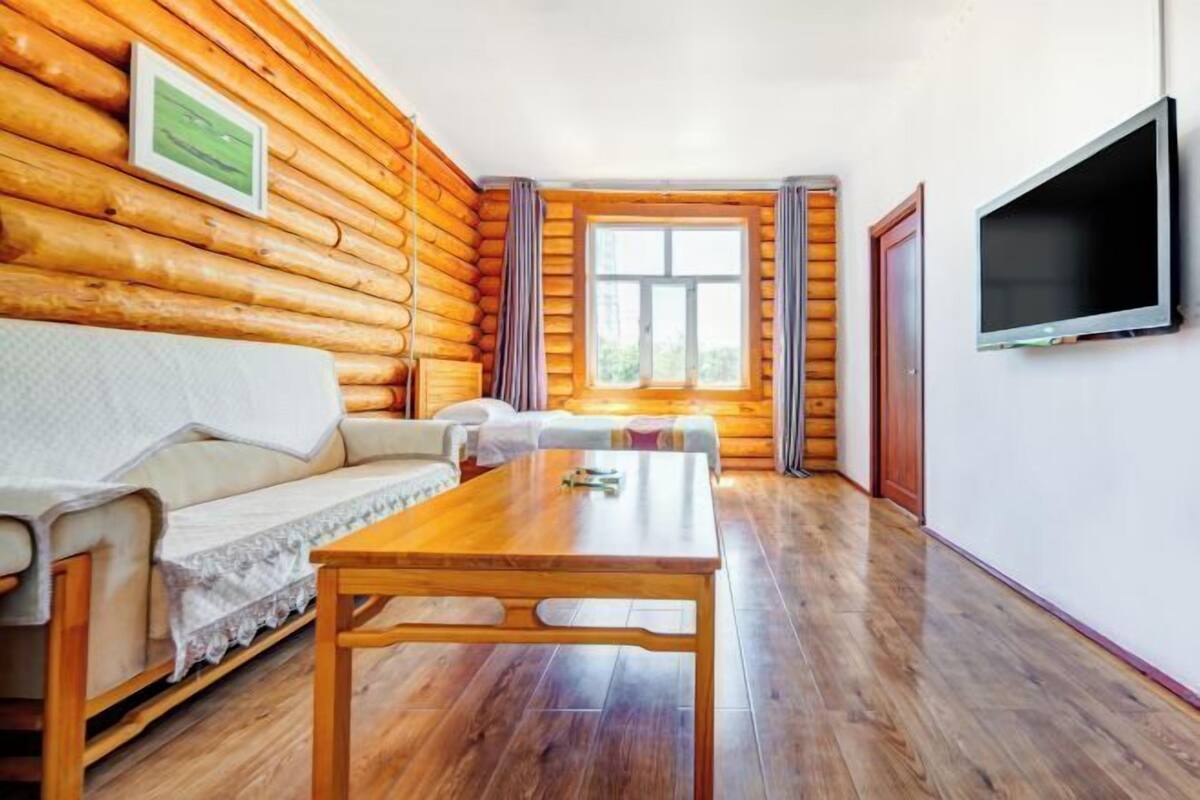 Una habitación tipo cabaña de troncos con piso de madera, un sofá, una mesa, un televisor y una ventana con cortinas.