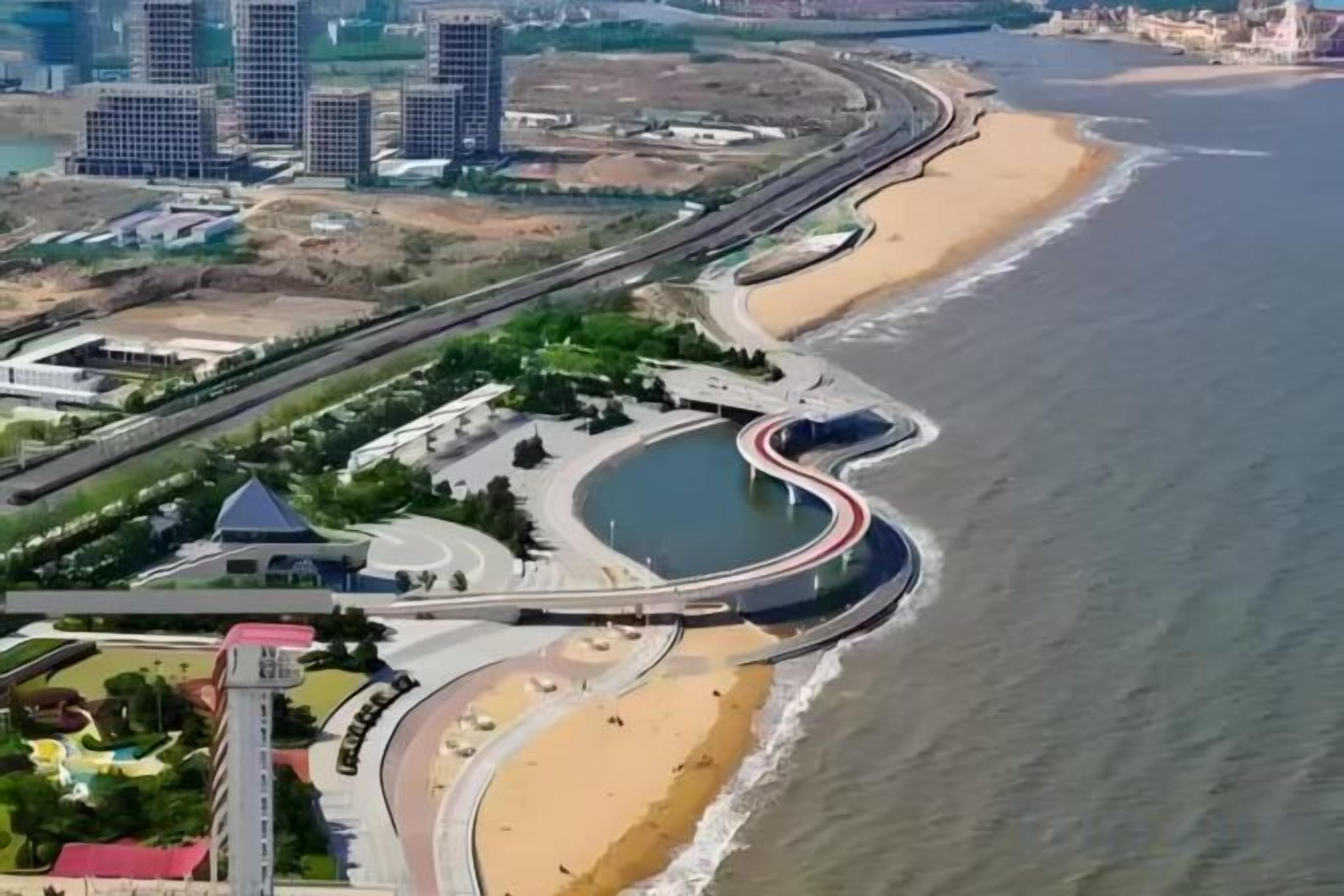An aerial view of a coastal area with a curved road, a beach, and buildings in the background.