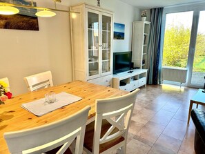 Ein Essbereich mit Holztisch und weißen Stühlen, einer Vitrine mit Glastüren, einem Fernseher auf einem Schrank und Blick auf Grün hinter einem großen Fenster.