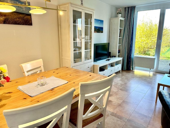 Un coin repas avec une table en bois et des chaises blanches, un meuble avec des portes en verre, un téléviseur posé sur un meuble, et une vue sur la verdure à travers une grande fenêtre.