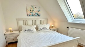 A neatly made bed with white bedding and pillows, a bedside table with a lamp, a painting on the wall, and a window with a view of trees.