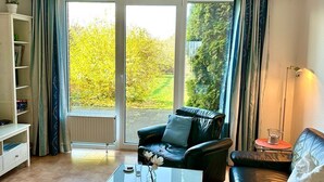 A living room with a black leather sofa, a glass coffee table, and a bookshelf. There is a sliding glass door leading to a garden.