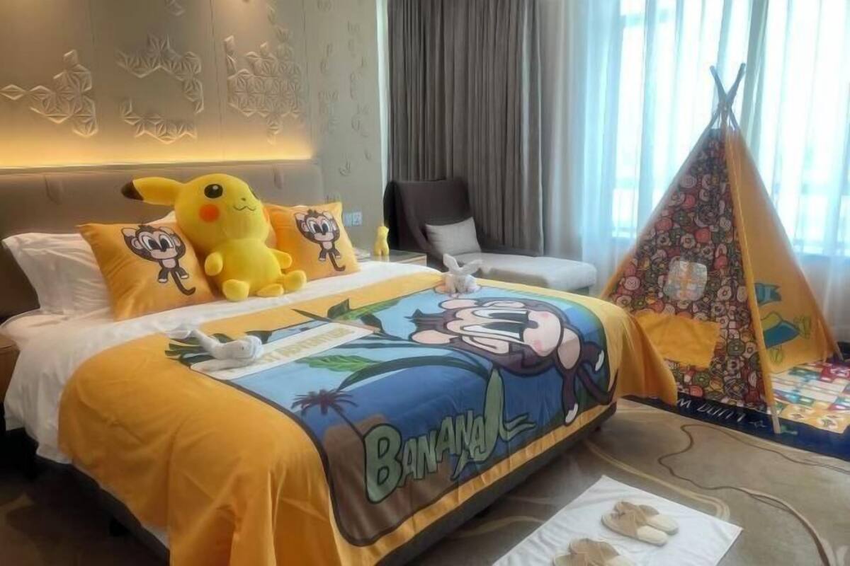 Hotellihuoneessa on sänky, jossa on banaaniteemalla koristeltu peitto, pehmeä Pikachu-pehmolelu ja sarjakuvahahmon kuva koristeltu tyyny. Huoneen nurkassa näkyy telttaa muistuttava rakenne.