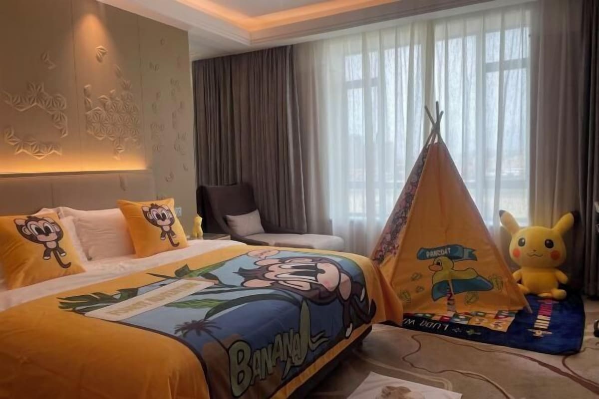 A hotel room with a bed featuring a banana-themed bedspread, a pillow with a monkey design, a chair, a window with curtains, and a small tent with a Pikachu plush toy.