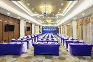 Une salle de conférence avec des rang ées de tables violettes, un écran de projection et une scène à l’avant.