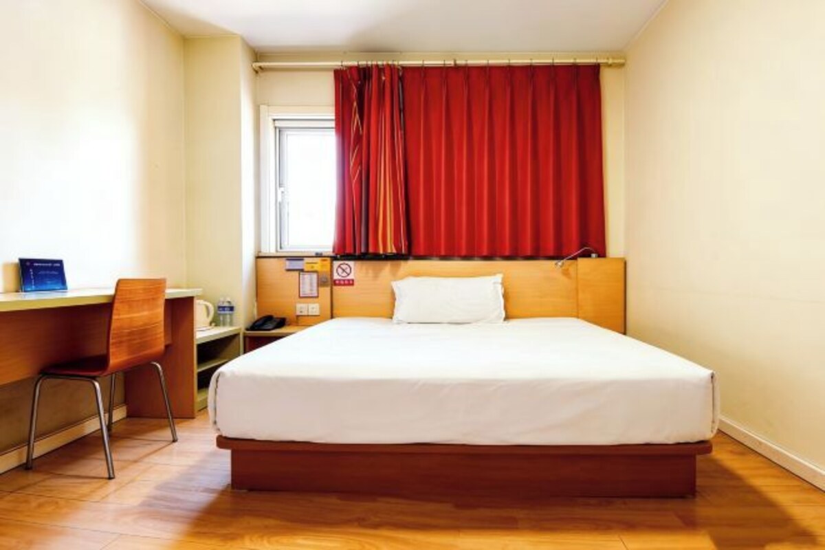 Una camera d'albergo con un letto, una scrivania, una sedia e una finestra con tende rosse.