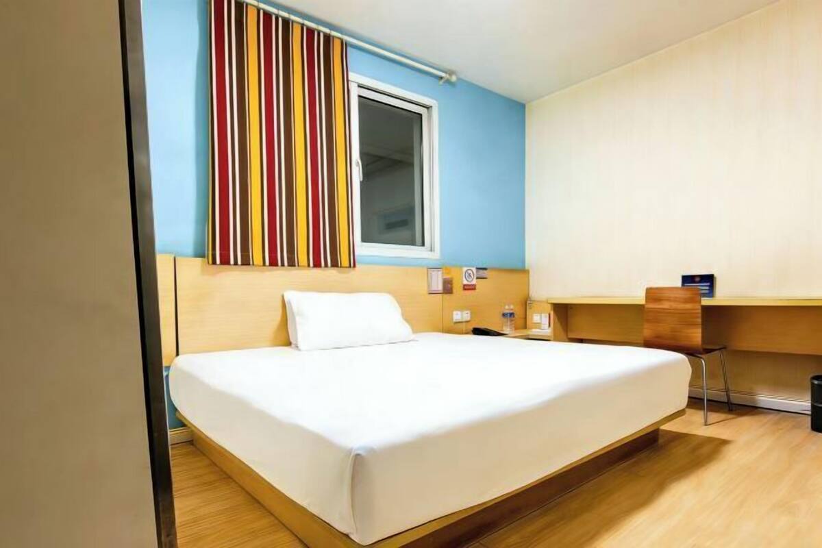 Une chambre d’hôtel avec un lit, un bureau, une chaise et une fenêtre avec un rideau à rayures.