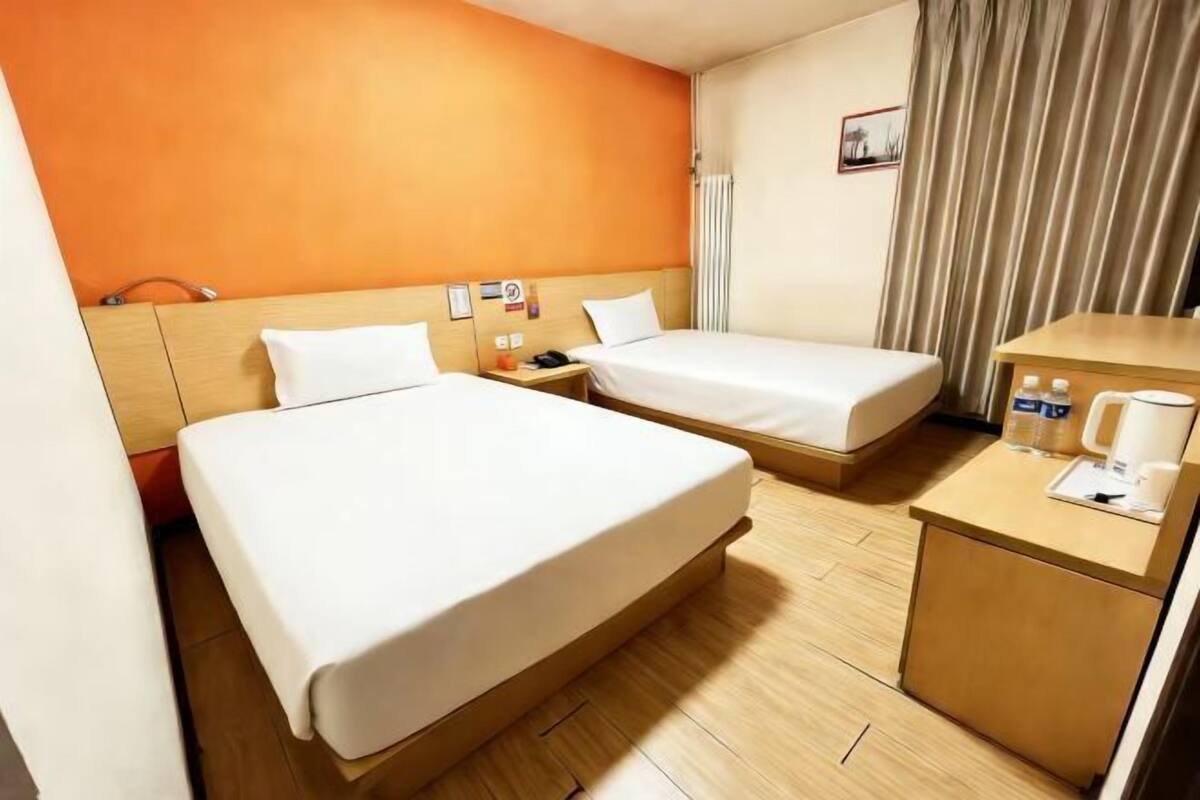 Une chambre d’hôtel avec deux lits, une tête de lit en bois, un petit bureau avec une lampe et un rideau.