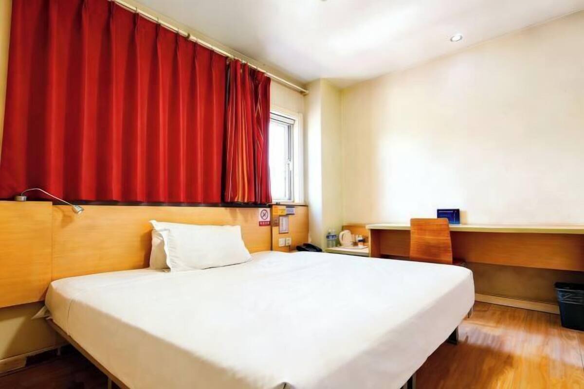 Une chambre d’hôtel avec un lit, un bureau, une chaise et une fenêtre avec des rideaux rouges.