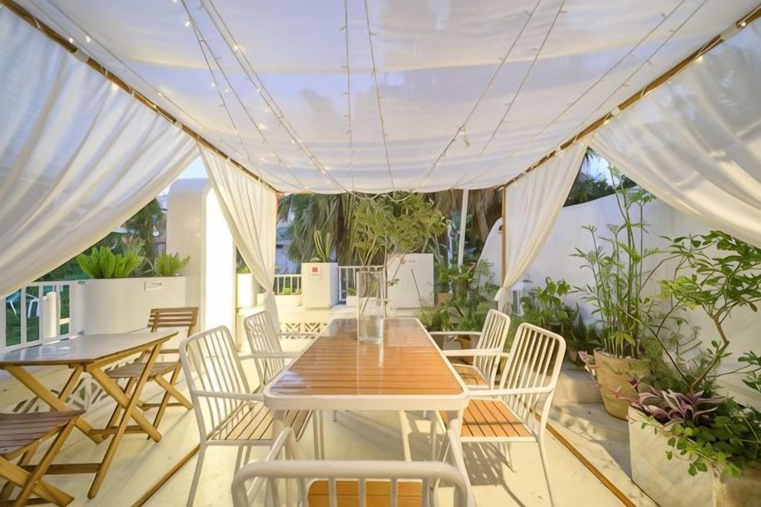 An outdoor dining area with a long wooden table, white chairs, and a white canopy.