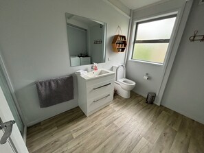 A modern bathroom with a white sink, toilet, and mirror. There is a window providing natural light and a small shelf with toiletries. - Otahu Retreat (Whangamata)