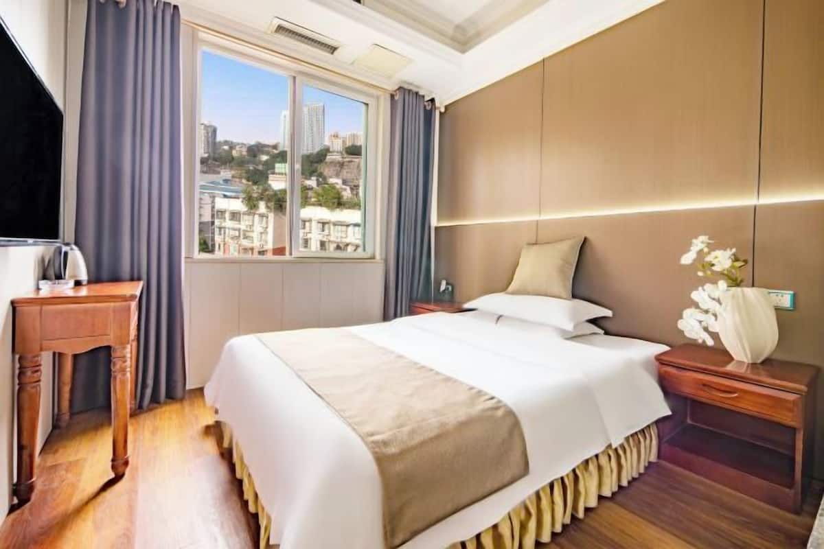 Une chambre d’hôtel avec un grand lit, une table de chevet en bois, un bureau, une fenêtre avec des rideaux et une vue sur les bâtiments.