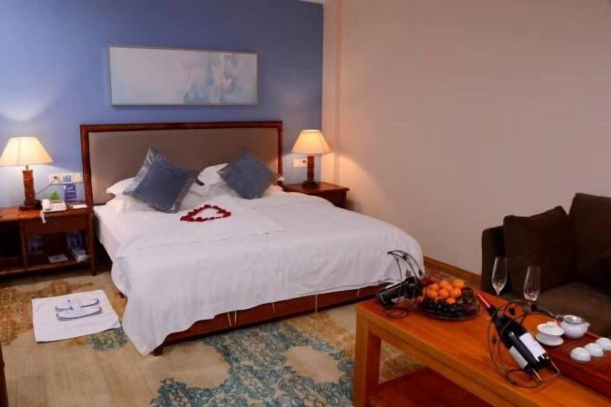 A hotel room with a bed, two bedside lamps, a nightstand, a wooden table with a fruit bowl and wine, and a painting on the wall.