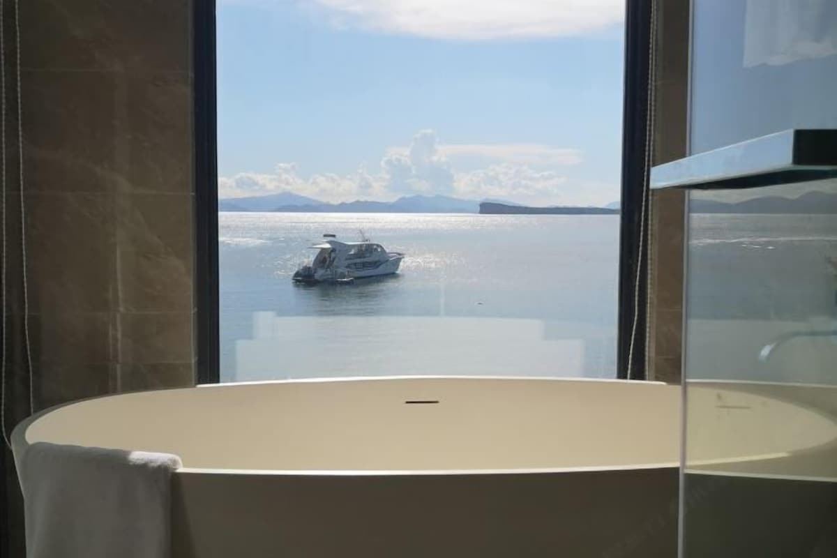 A bathroom with a large bathtub, a window with a view of a calm body of water and a boat.
