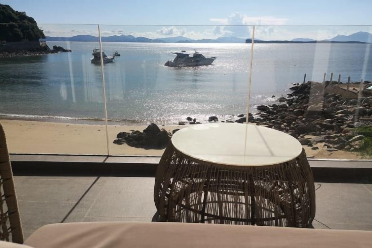 A balcony with a wicker chair, a view of calm water, boats, and a rocky shoreline.