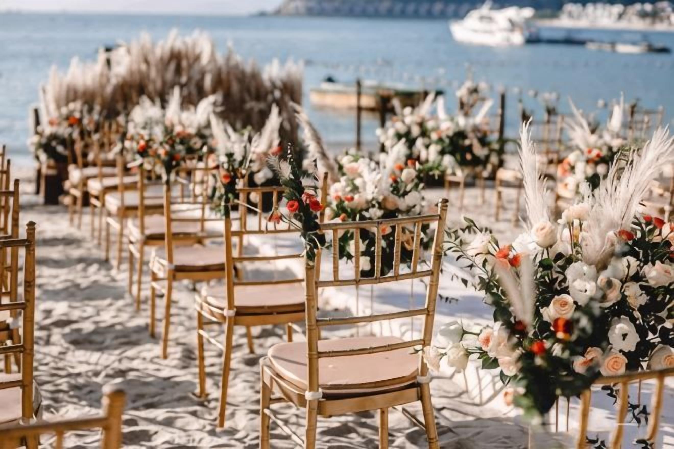 Rows of wooden chairs with floral arrangements, set on a sandy beach with a boat in the background.