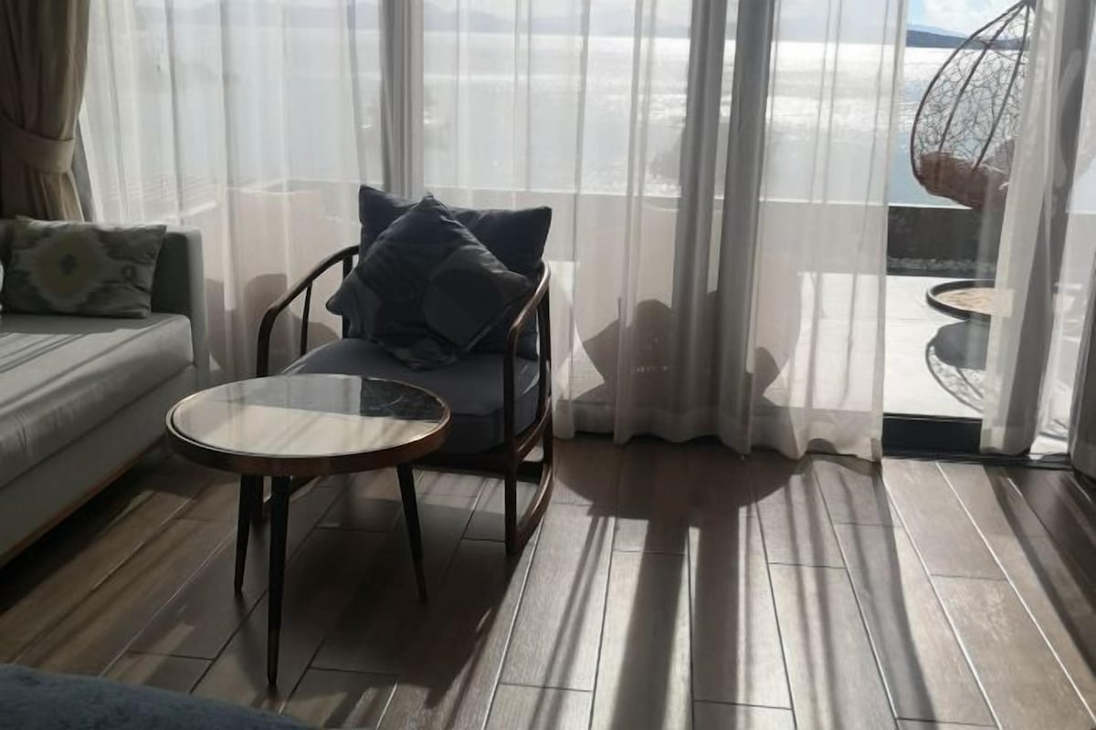 A room with a sofa, a chair with cushions, a small table, and a view through sheer curtains.