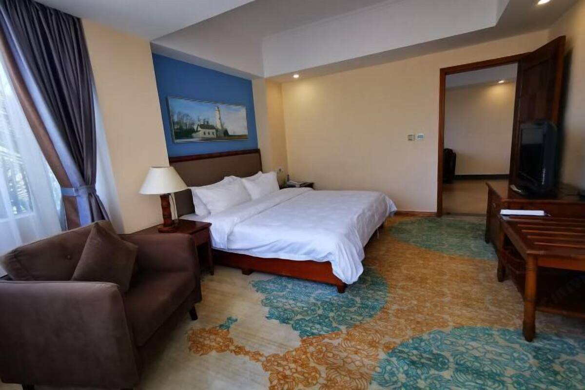 A hotel room with a bed, a desk, a chair, a TV, and a painting on the wall.