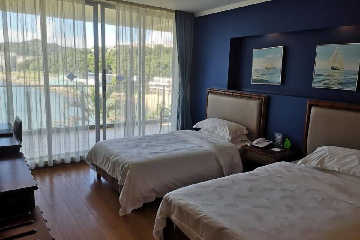 A hotel room with two beds, a desk, a chair, a TV, and a view of the outdoors.