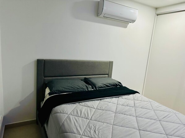 A bed with a quilted white duvet and two pillows, a grey headboard, and a white air conditioning unit mounted on the wall. - Unit 6 house (Oranjestad)