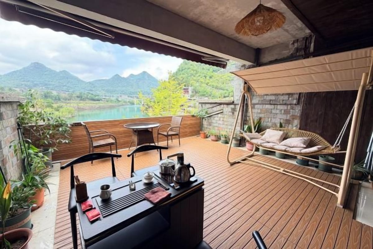 Una terraza de madera con una mesa y sillas, una hamaca colgante y vistas a un lago y montañas.