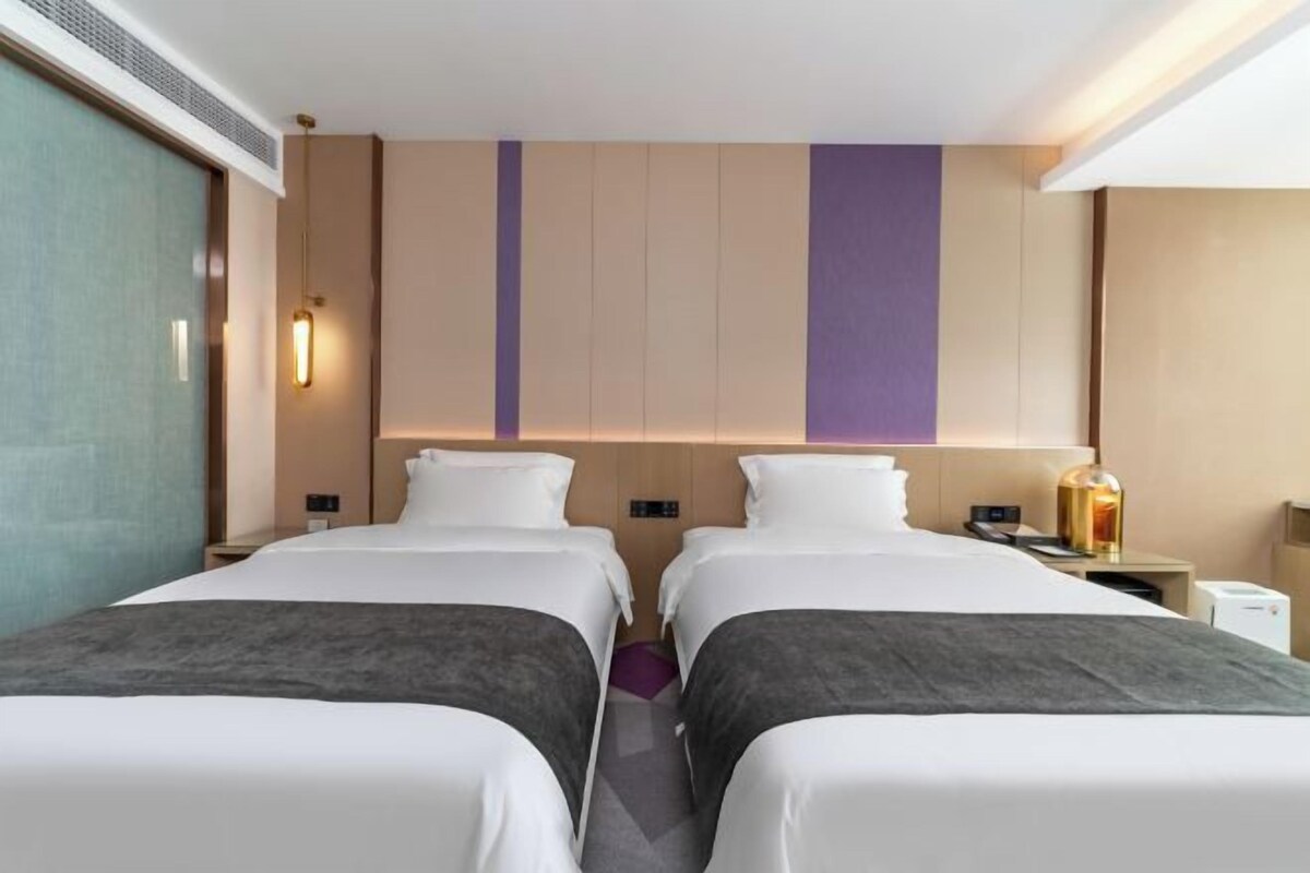 A hotel room with two beds, a nightstand, a lamp, and a wall-mounted control panel.