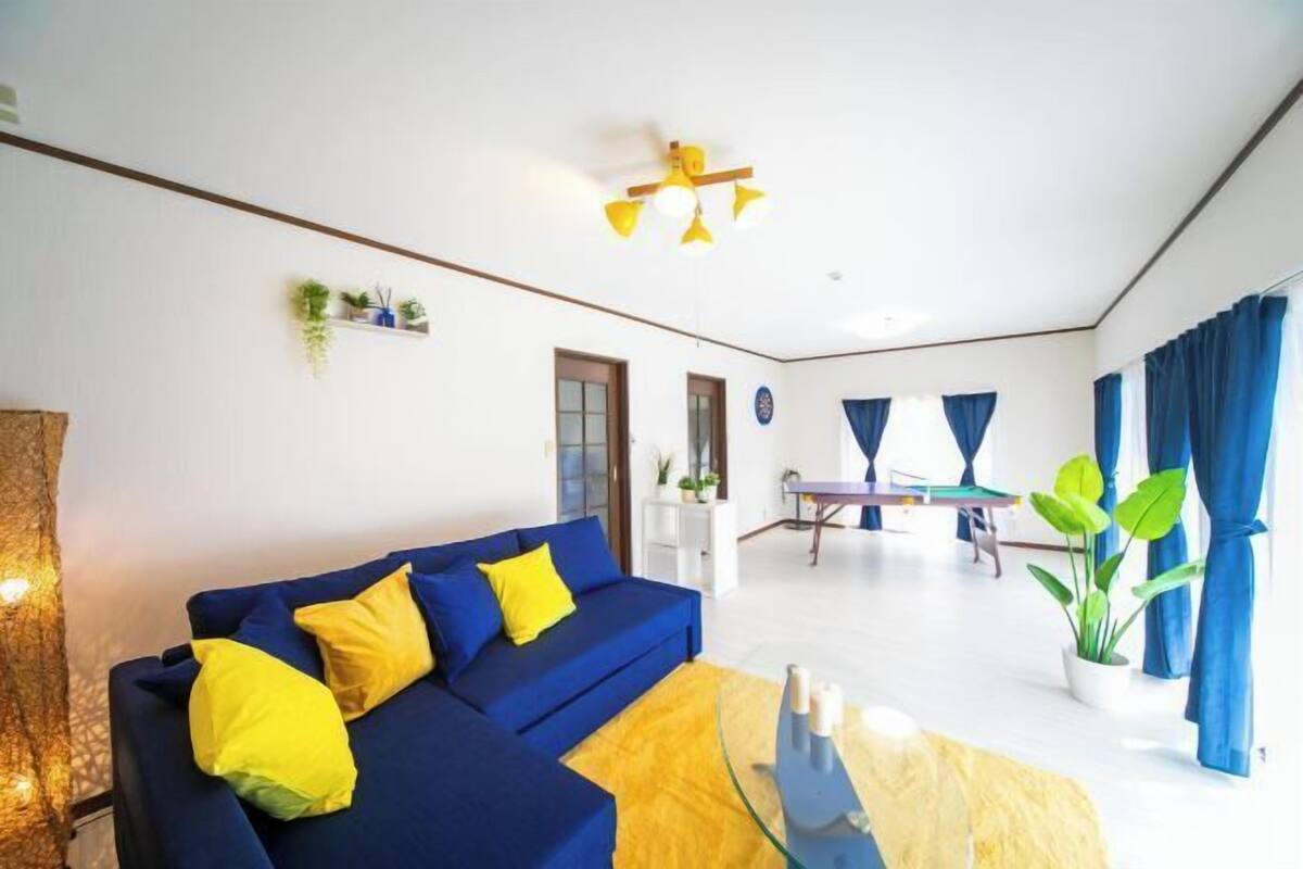Uma sala de estar com um sofá azul, almofadas amarelas, uma mesa de centro de vidro, um ventilador de teto e uma área de jantar com mesa e cadeiras.