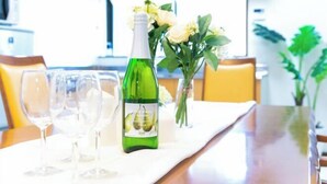 Mesa de comedor dispuesta con copas de vino, una botella y un florero en una habitación luminosa con sillas amarillas y una cocina al fondo.