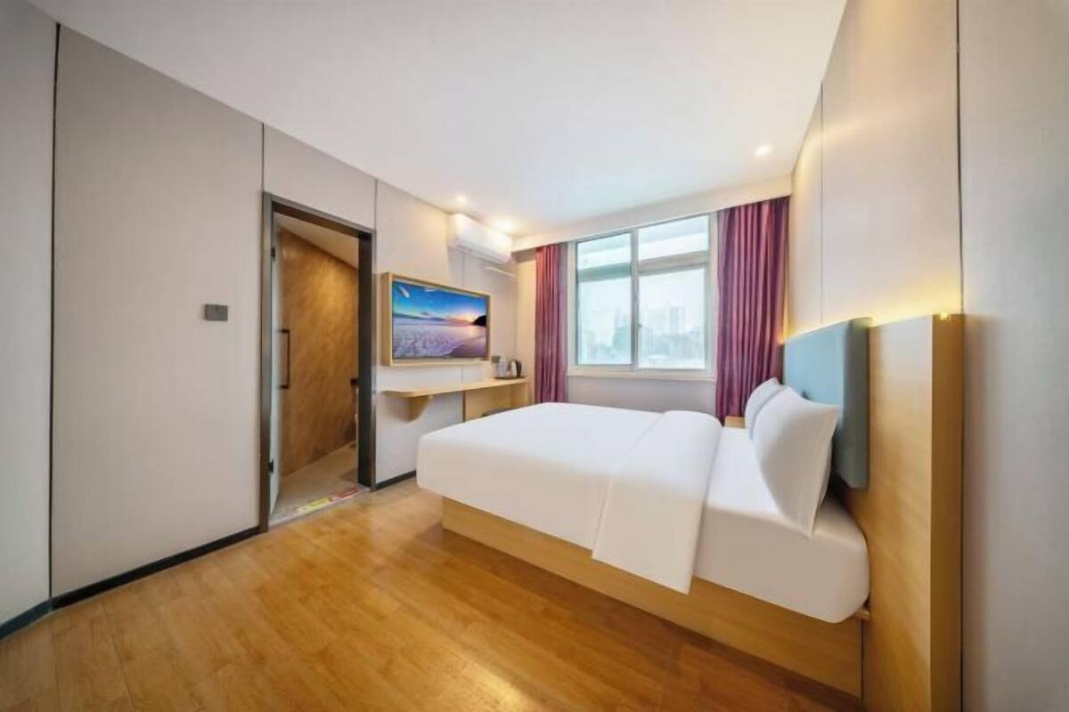 A hotel room with a bed, a TV, a window with curtains, and a cityscape view.