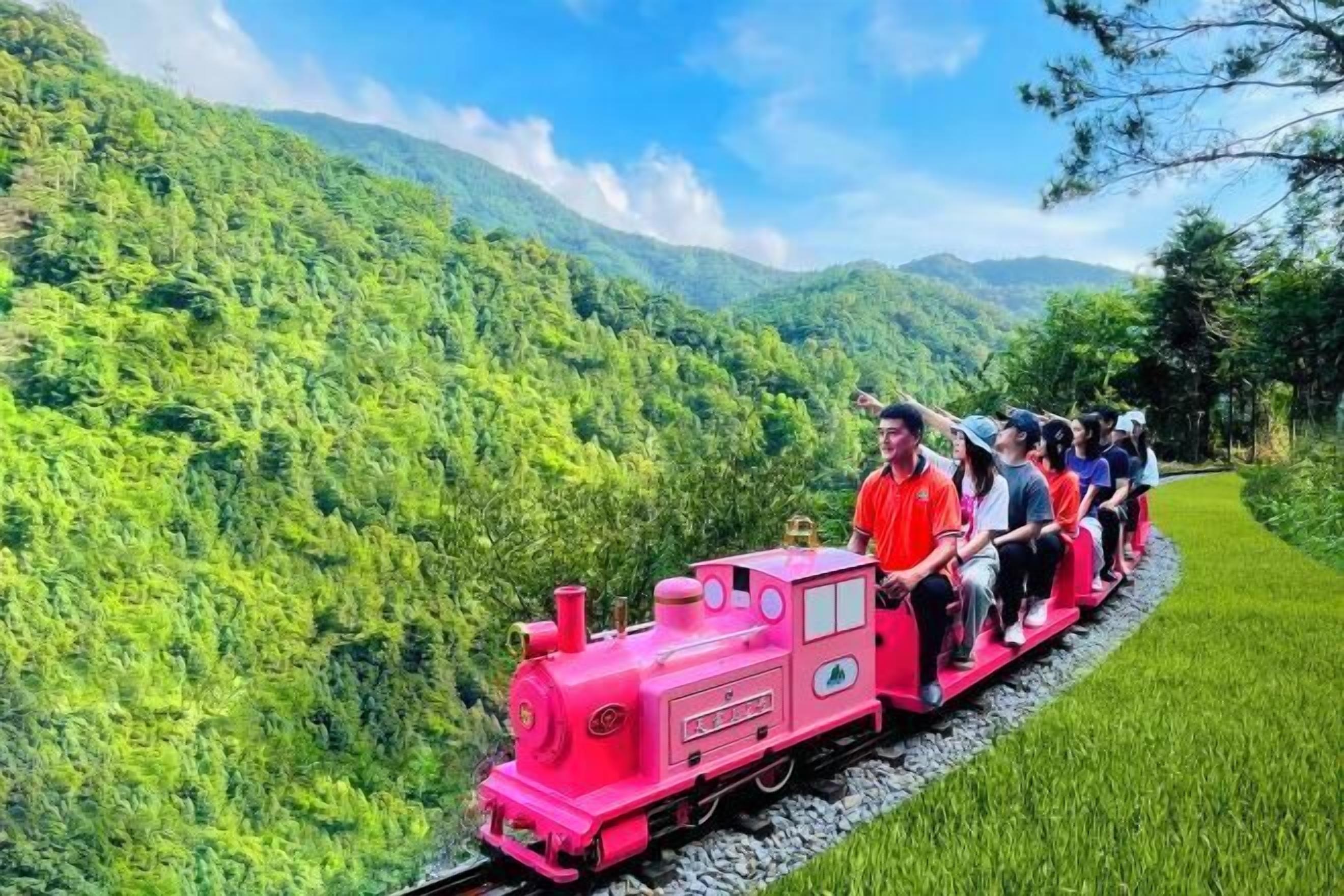 A pink train with passengers on a track surrounded by lush greenery.
