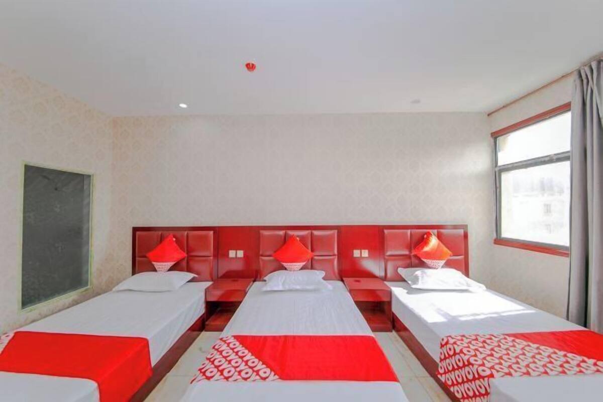 A hotel room with three beds, a large window, and a red headboard.