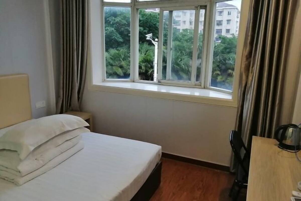 A hotel room with a bed, a window with curtains, a wooden floor, and a desk with a kettle.