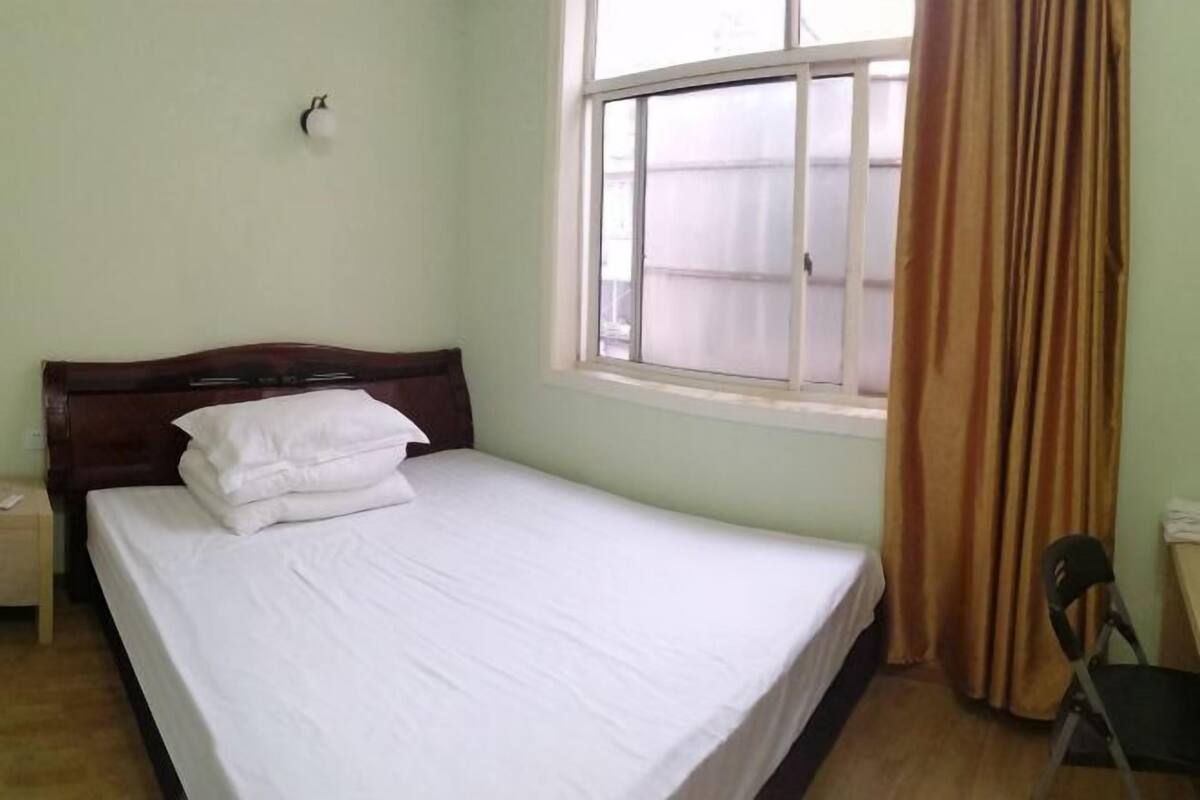 A single bed with white bedding, a wooden headboard, a window with curtains, and a small table with a chair.