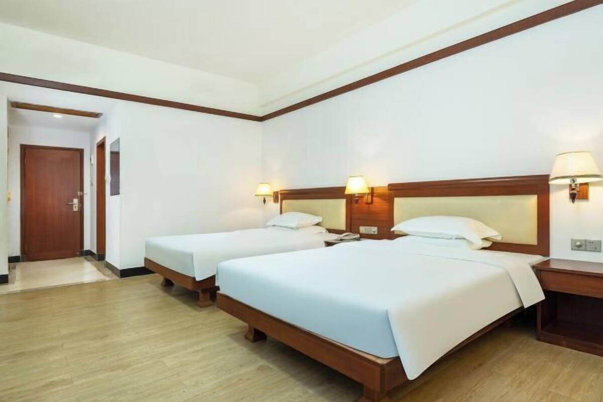 A hotel room with two single beds, wooden headboards, bedside lamps, a wooden nightstand, and a wooden door.