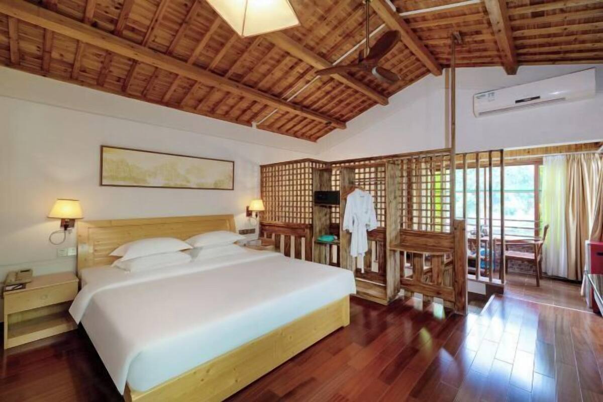 A room with a wooden ceiling, a bed with white linens, a nightstand, a wall-mounted lamp, and a window with curtains.