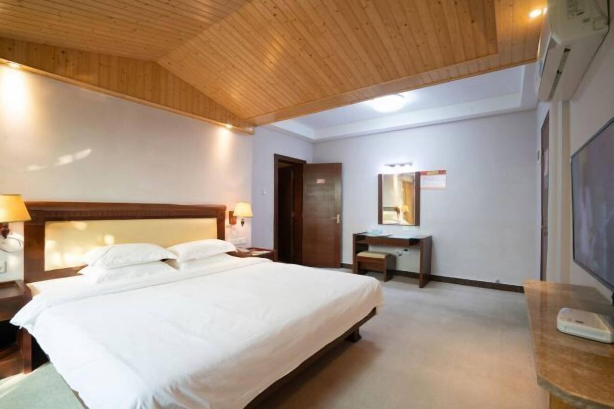 A hotel room with a wooden ceiling, a bed, a bedside table with a lamp, a mirror, a desk, and a television.